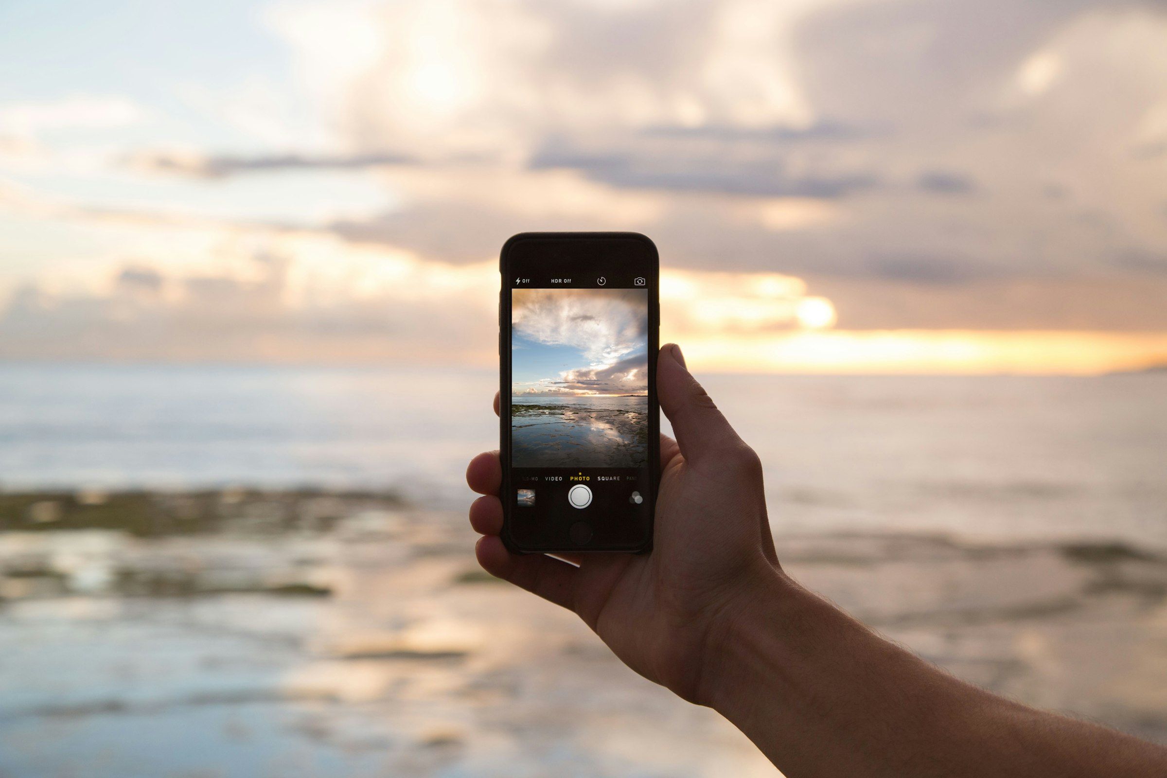 A hand holding a smartphone in front of the sea at sunset